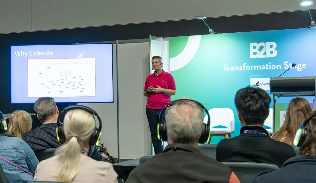Speaker in a pink polo presenting on stage beside a LinkedIn slide, addressing an audience at a conference.