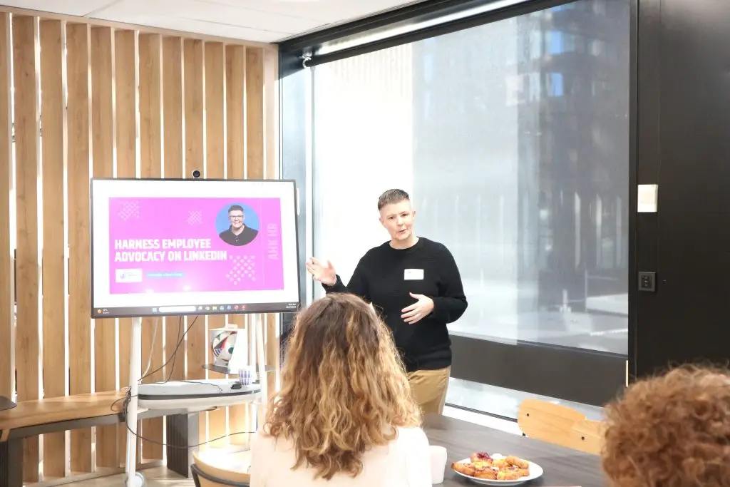 Speaker presenting a slide titled 'Harness Employee Advocacy on LinkedIn' to a small audience in a bright room with a pink graphics slide behind him.